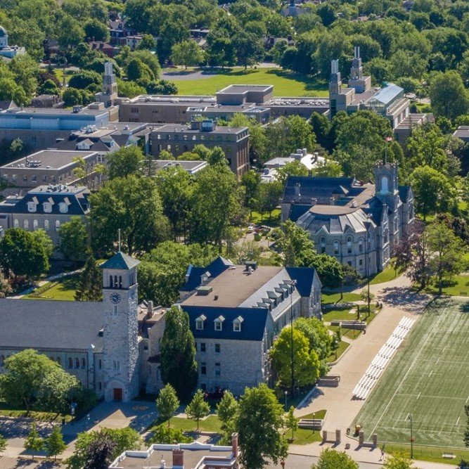 Aerial shot of Queen's campus with a focus on the Grant Hall clocktower. The weather is sunny and the trees are green and vibrant.